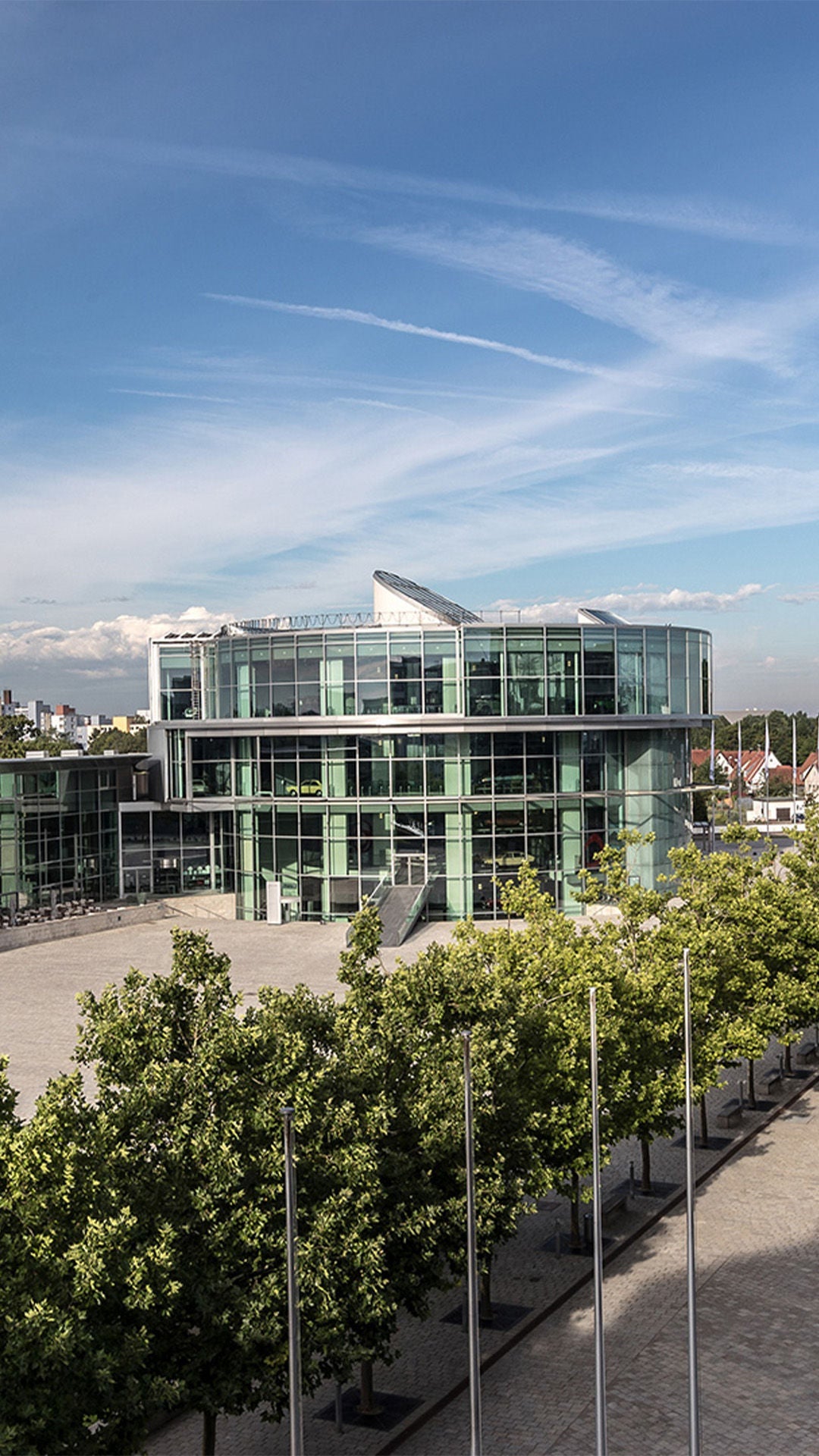 Blick auf moderne Gebäude mit Glasfassaden, breitem Vorplatz und begrüntem Bereich bei Tageslicht.