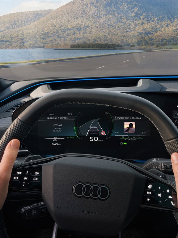 Zwei Hände halten das Lenkrad eines Audi Q6 Sportback e-tron, digitale Anzeigen und Displays zeigen Geschwindigkeit und Musik.