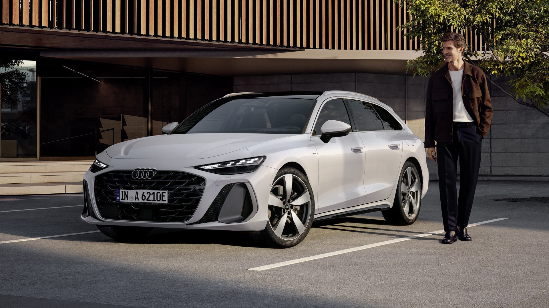 A6 Avant e-hybrid med sluttande frontprofil på en parkeringsplats framför en stor byggnad med träpanel. En medelålders man står bredvid fordonet.