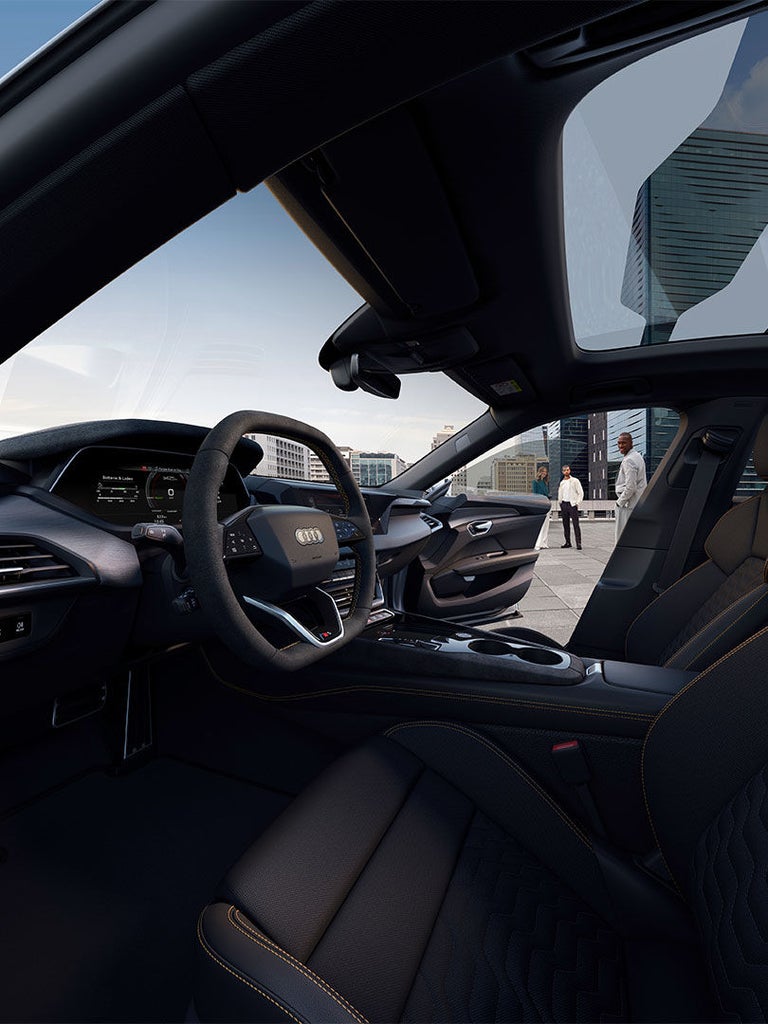 Innenraum des Audi S e-tron GT mit sportlichem Lenkrad, digitalen Anzeigen und hochwertigen Materialien. Durch das offene Fenster sind drei Personen auf einer Dachterrasse mit Stadtblick zu sehen.