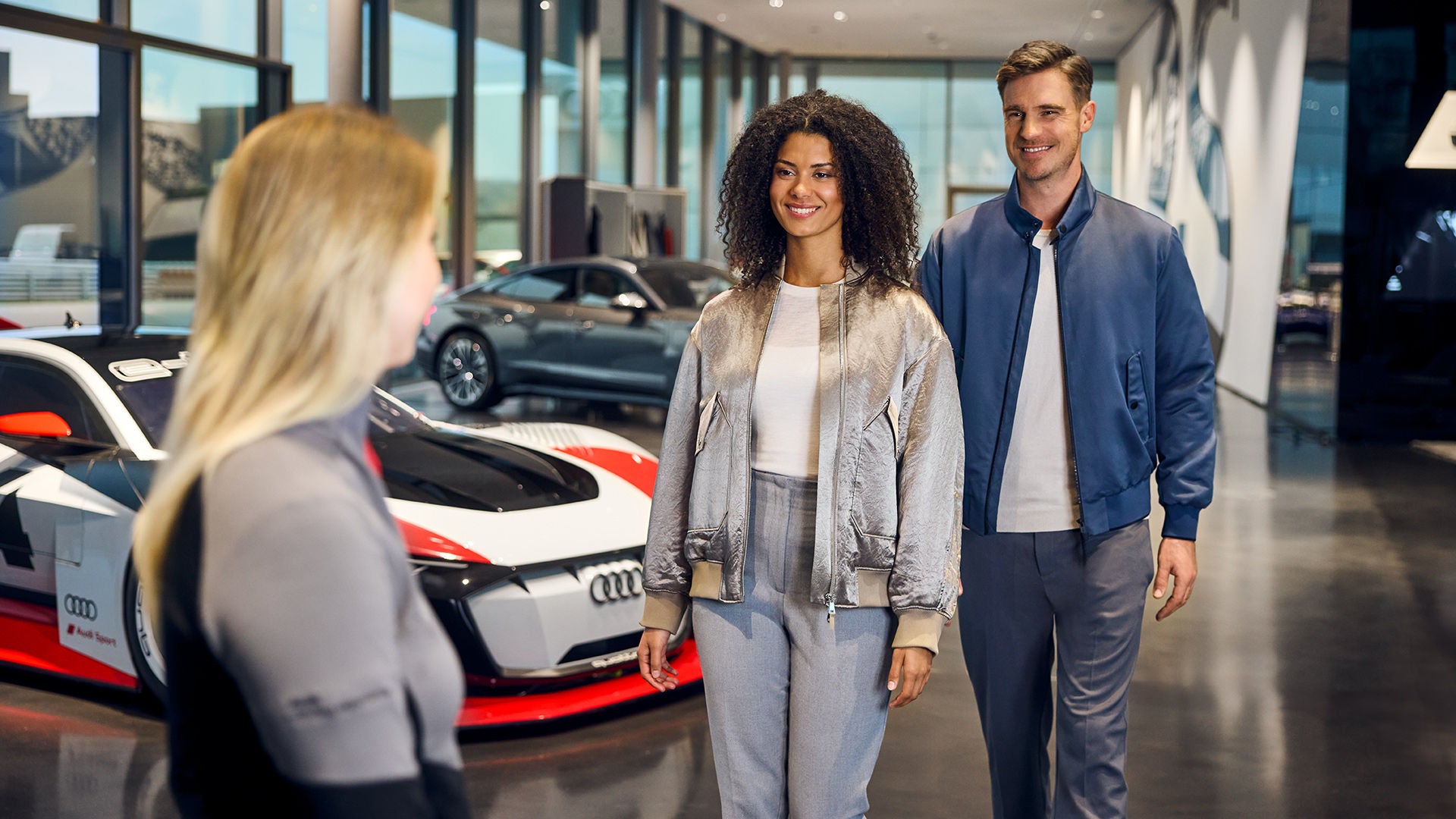 Zwei lächelnde Personen im Gespräch mit einer Mitarbeiterin in einem Audi Showroom mit Sportwagen im Hintergrund.