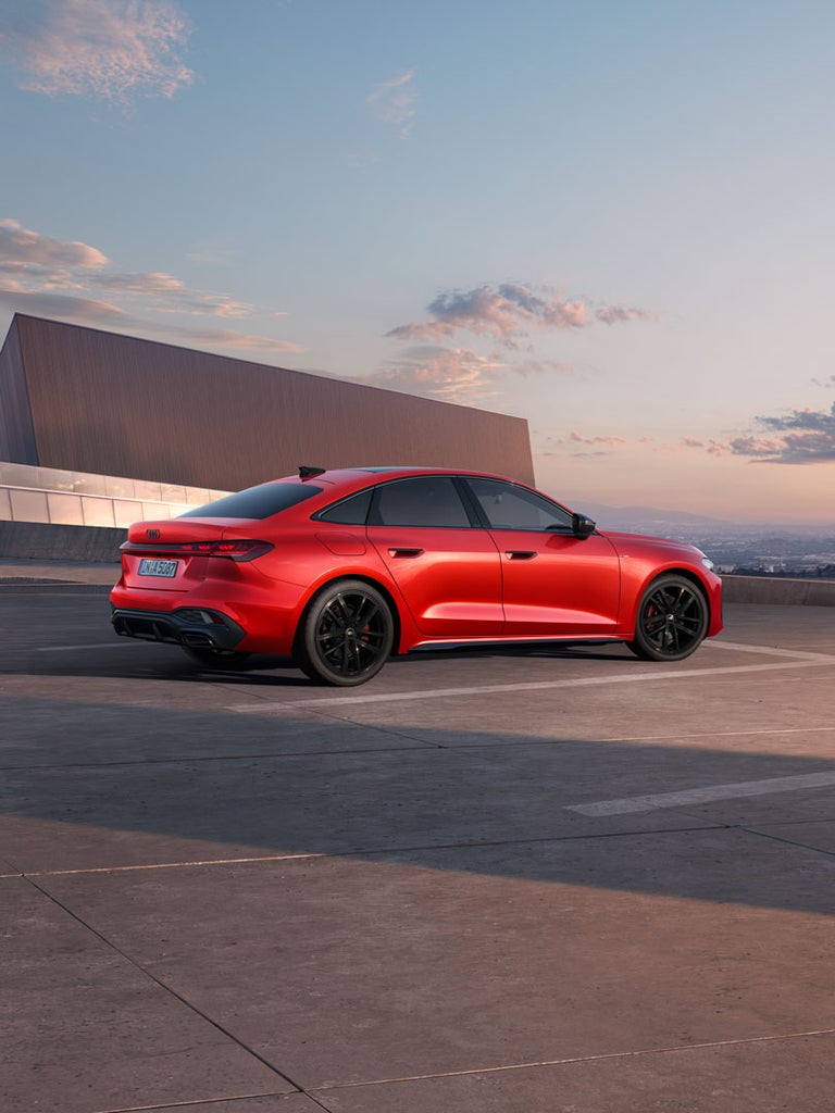 Rote Audi A5 Limousine von hinten seitlich, geparkt vor moderner Architektur bei Sonnenuntergang.