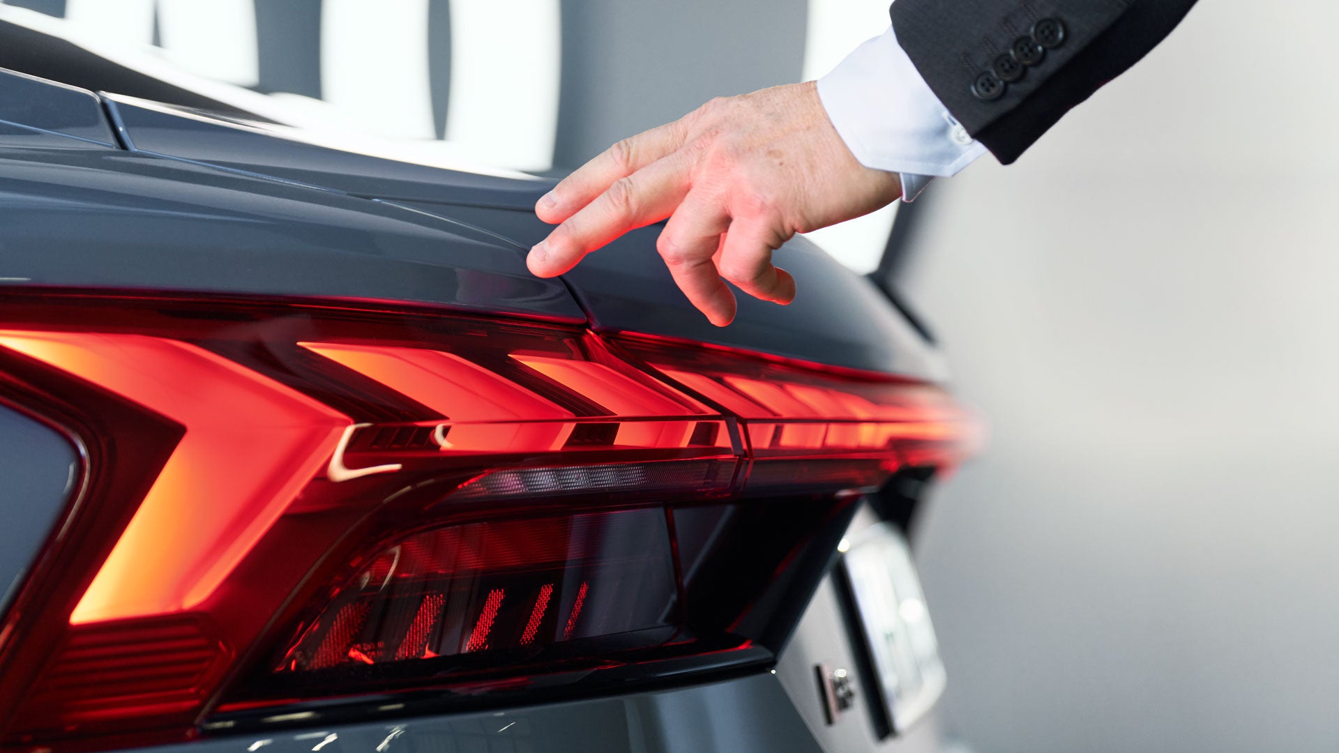 Hand eines Mannes zeigt auf das leuchtende LED-Rücklicht eines grauen Audi-Fahrzeugs im Innenraum eines Autohauses.