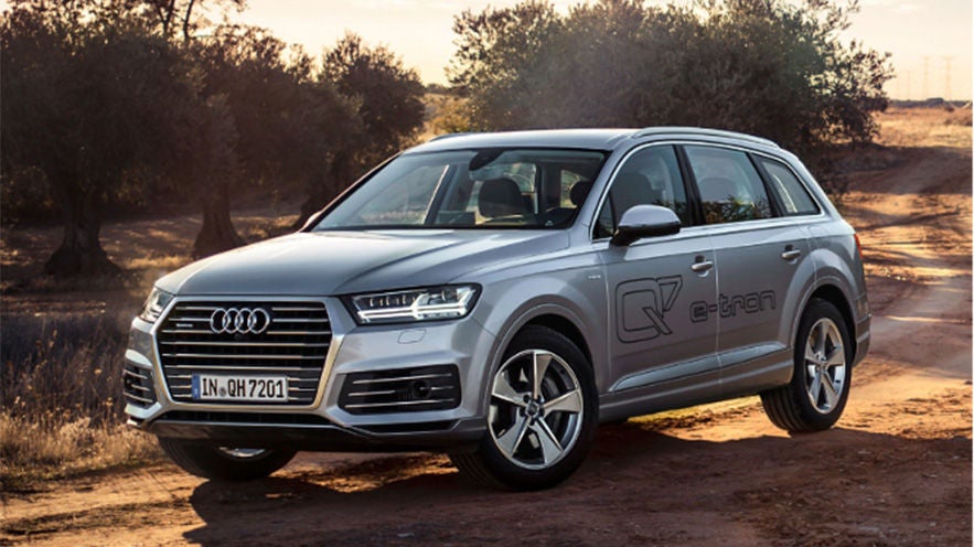 Silberner Audi Q7 e-tron steht auf einem Feldweg bei Sonnenuntergang, umgeben von Bäumen und trockenem Gelände.