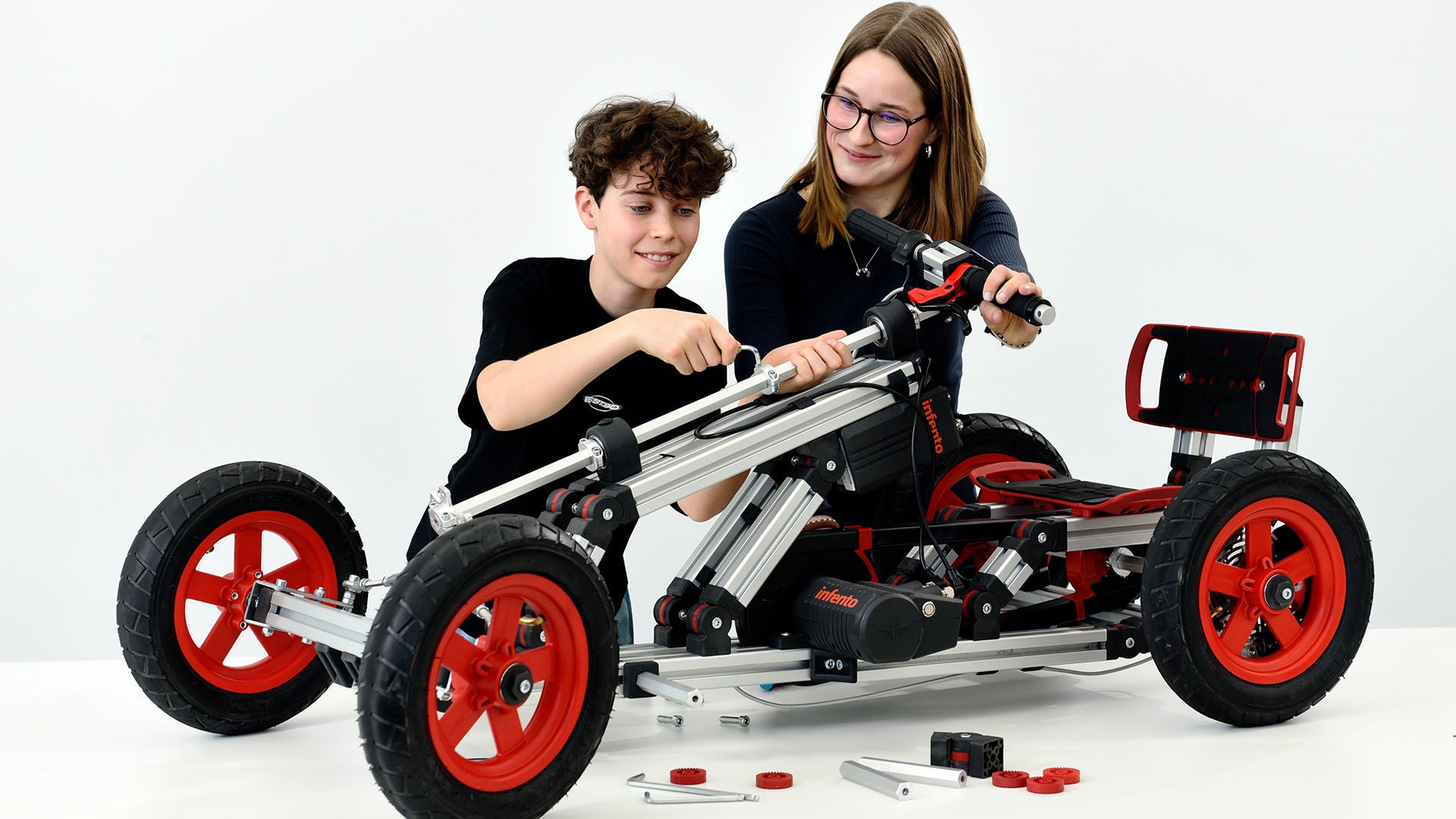 Zwei Jugendliche montieren ein vierrädriges Fahrzeugmodell mit roten Rädern auf weißem Hintergrund.