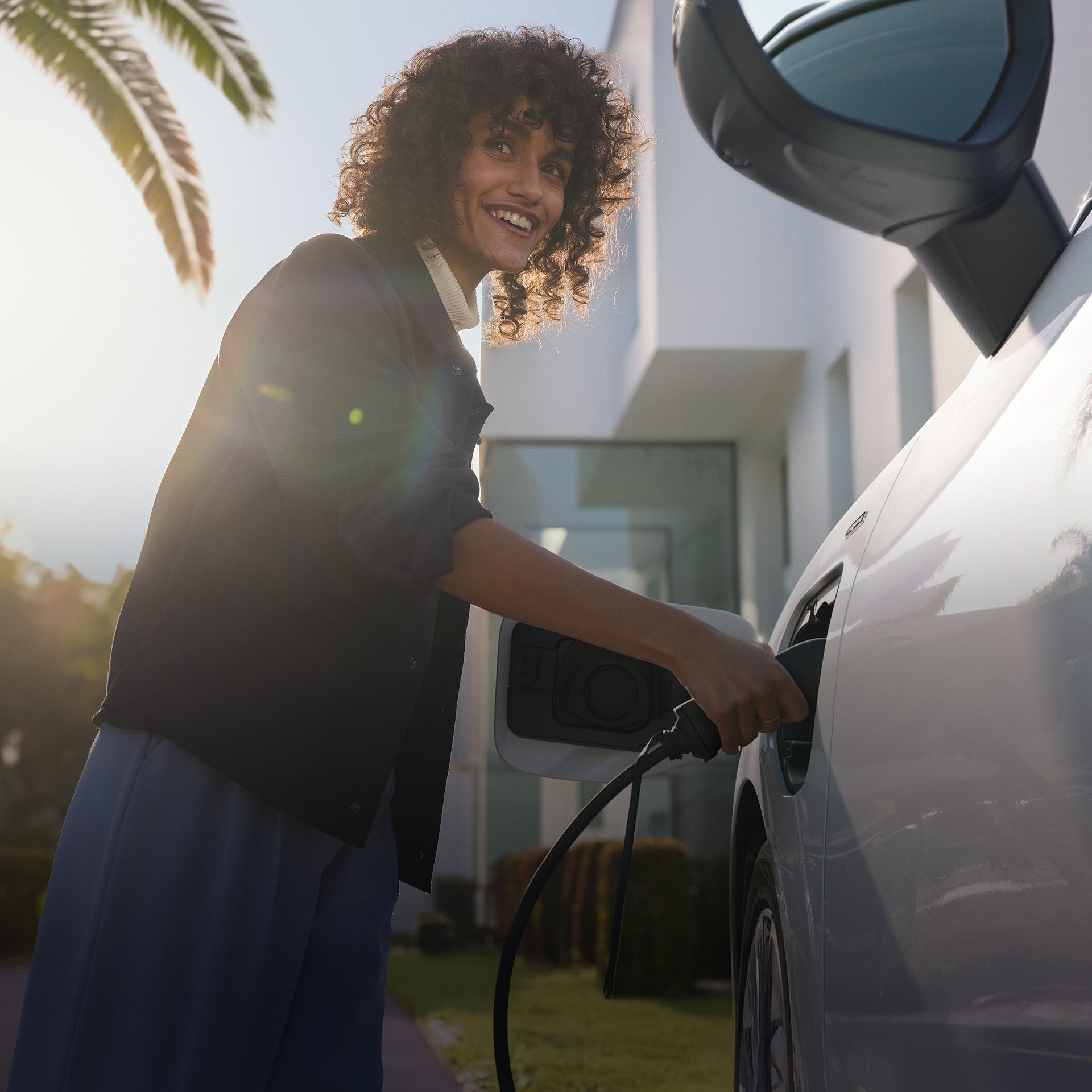 Frau lädt lächelnd ein weißes Elektroauto bei Sonnenschein vor einem modernen Gebäude mit Palmen.