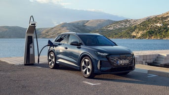 Audi-SUV steht an Ladesäule am Wasser; Ladekabel angeschlossen, Berge und Küstenlandschaft im Hintergrund.