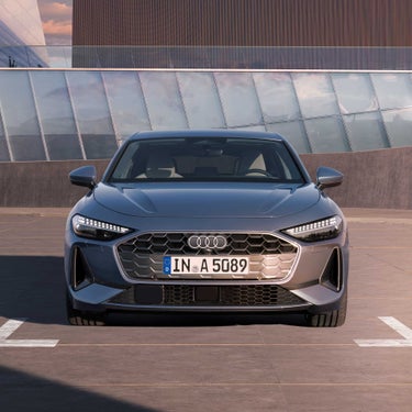 Frontansicht einer grauen Audi A5 Limousine mit markantem Kühlergrill und LED-Scheinwerfern vor moderner Glasfassade.