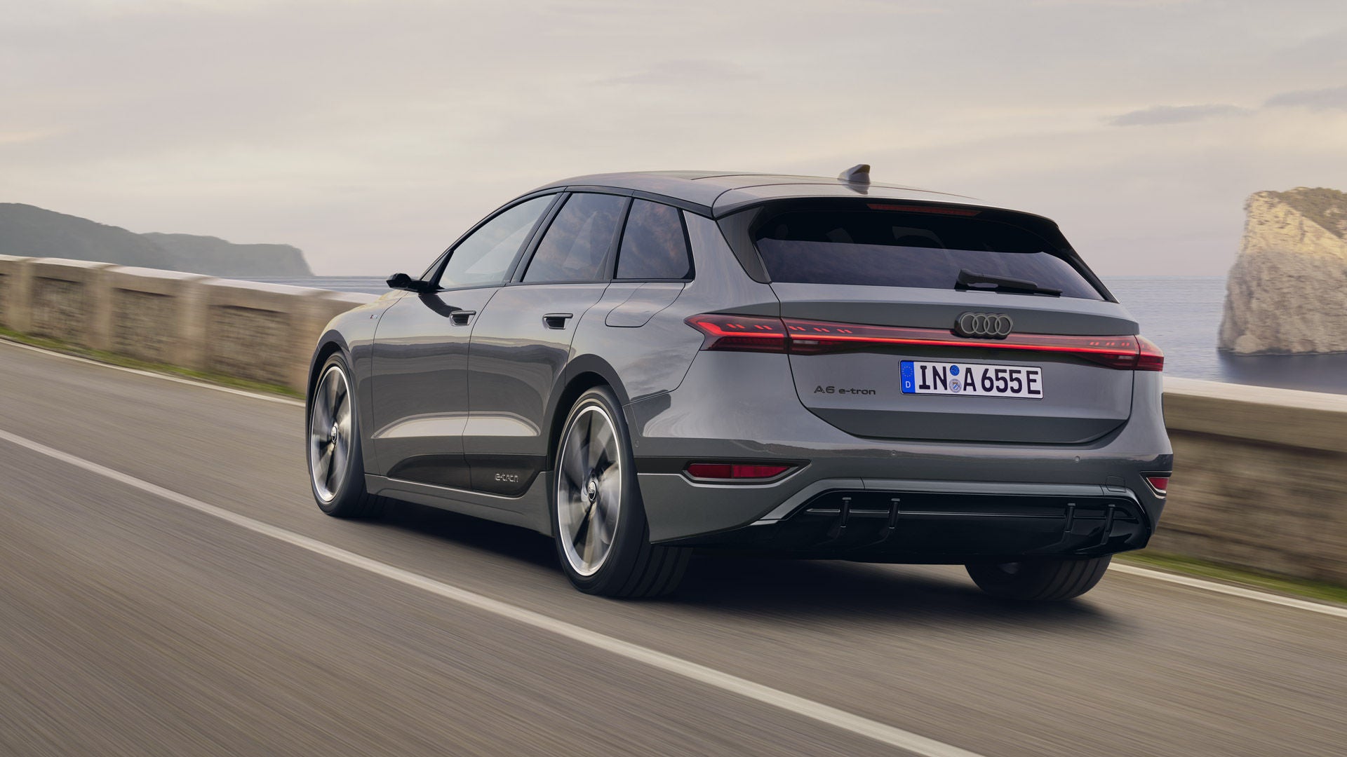 Schräge Heckaufnahme des A6 Avant e-tron, welcher auf einer Straße entlang des Meeres fährt. Im Hintergrund sind Felsen und Berge zu erkennen.&nbsp;