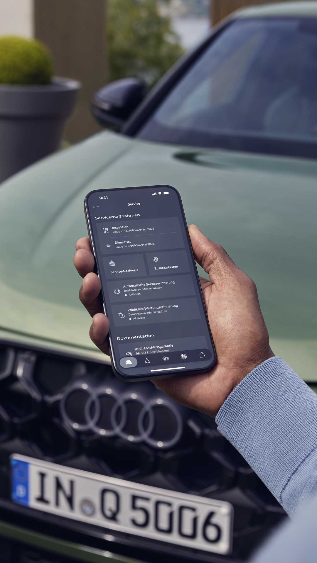 Die Hand eines Mannes ragt von unten ins Bild. Er hält ein Smartphone in der Hand, auf dessen Display Service-Funktionen angeboten werden. Im Hintergrund unscharf ein grüner Audi Q5.