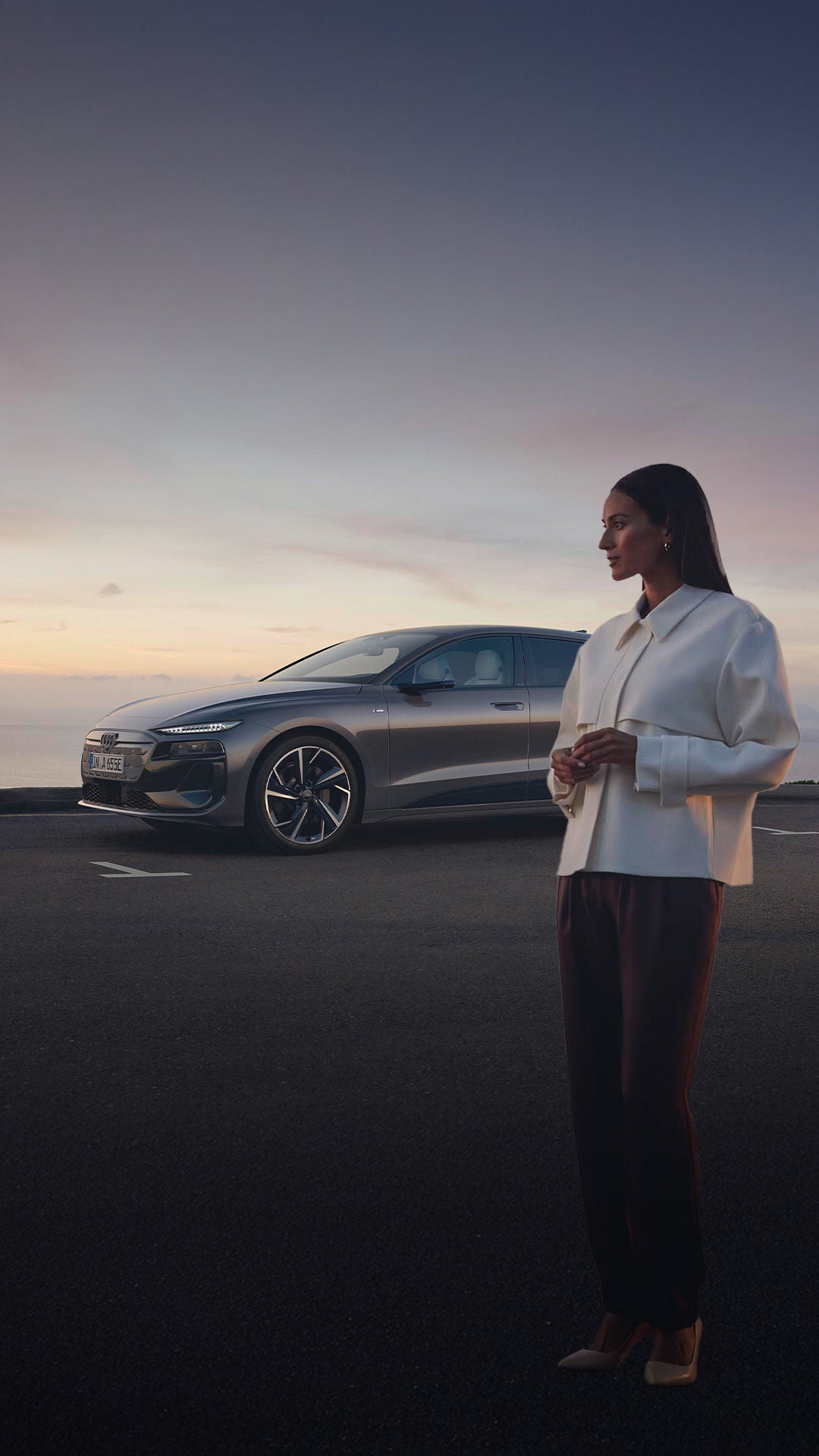 Grauer Audi A6 e-tron parkt vor Meer bei Sonnenuntergang, Frau in weißer Jacke steht daneben