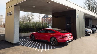 Rotes Audi-Elektroauto lädt tagsüber an einer überdachten Ladestation mit Audi-Logo und mehreren Ladepunkten.