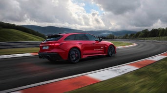 Roter Audi RS 5 Avant fährt auf Rennstrecke durch Kurve, bewölkter Himmel und grüne Hügel im Hintergrund.