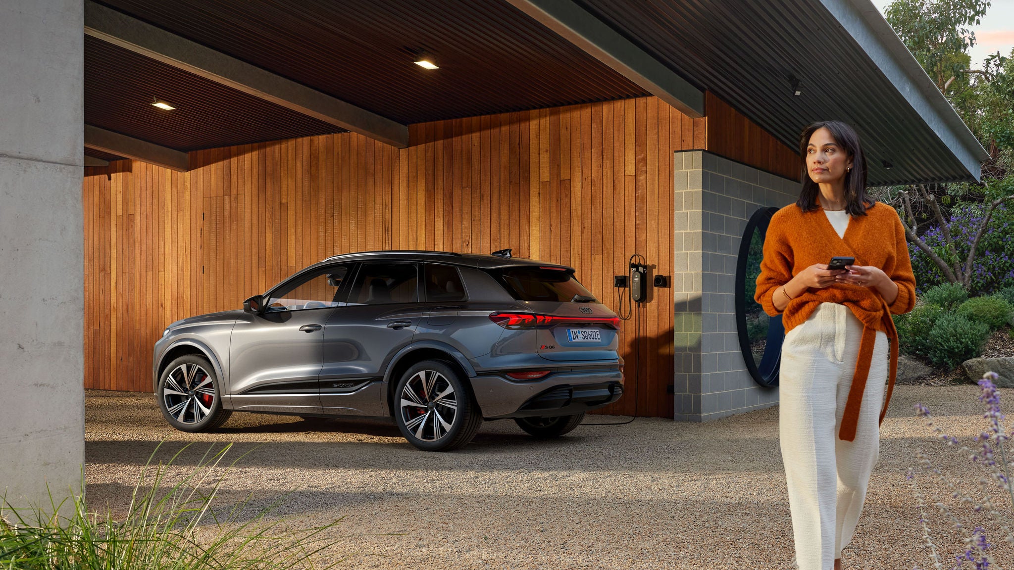 Eine Frau steht mit Smartphone neben einem silbernen Audi Q6 e-tron, der an einer Hausladestation lädt.