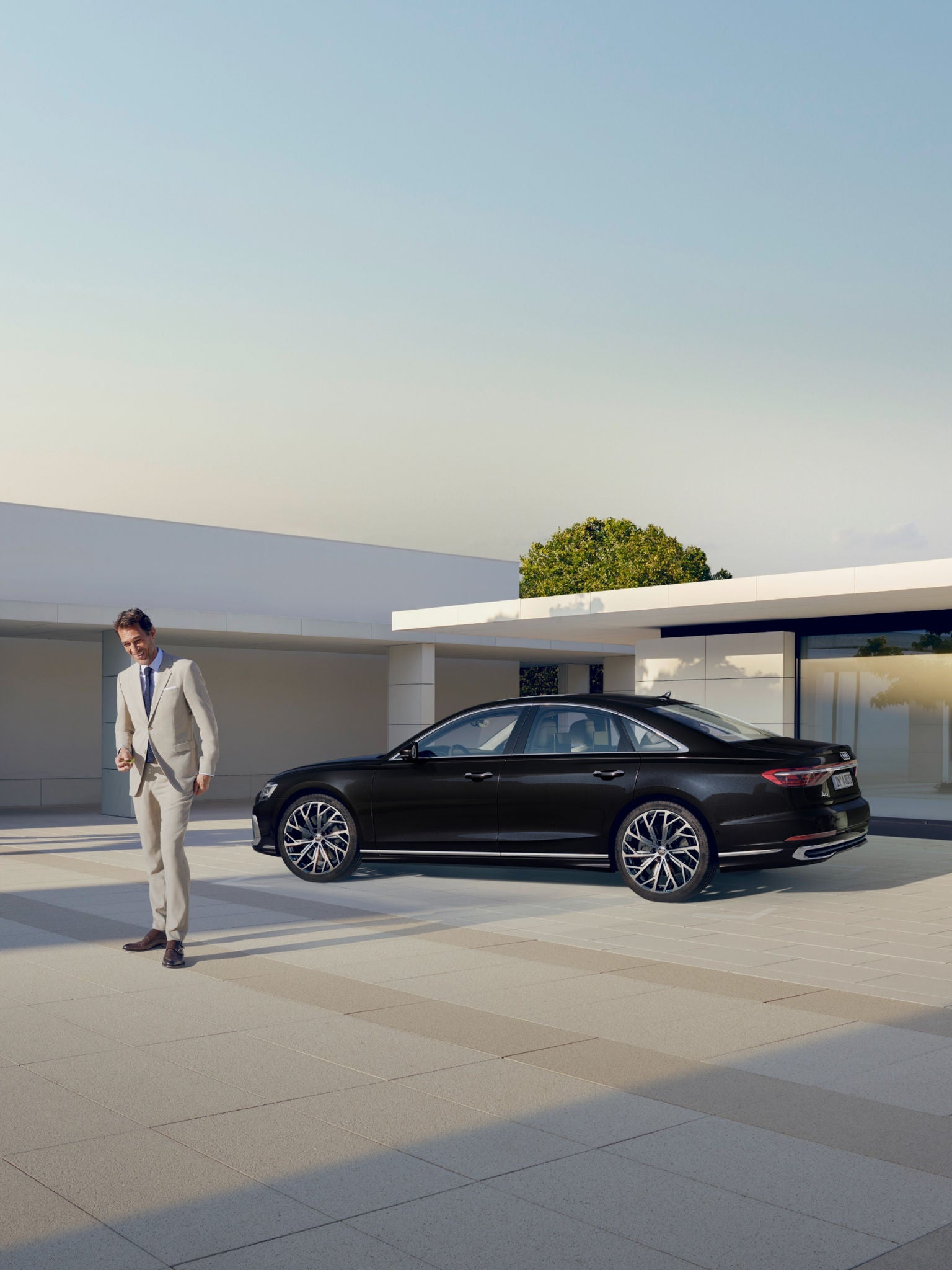 A person in a white suit walking towards a black car parked outside a modern building.