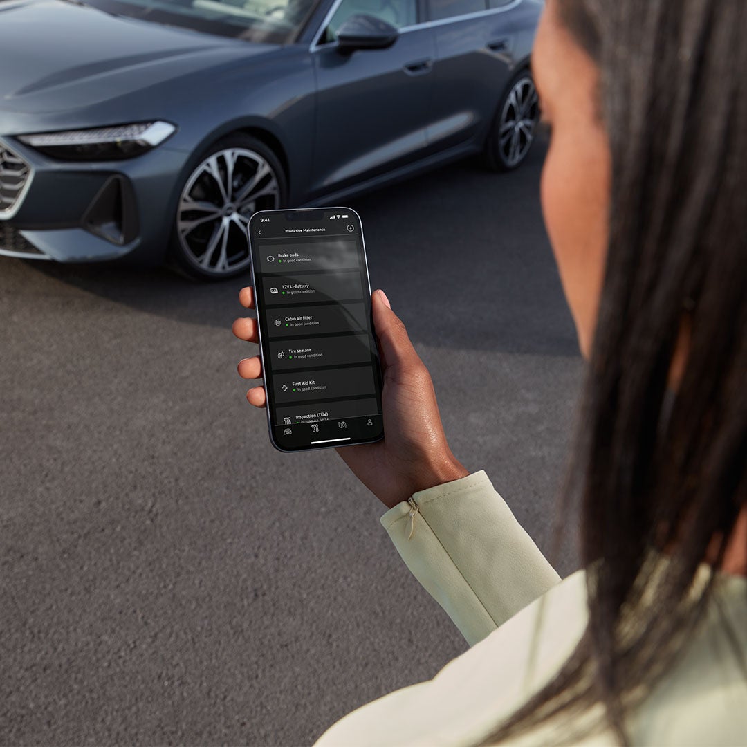 Une femme aux cheveux foncés tient un téléphone portable dans sa main, sur l'écran duquel on peut voir l'interface de l'application Audi. Une Audi sombre est garée à l'arrière-plan.