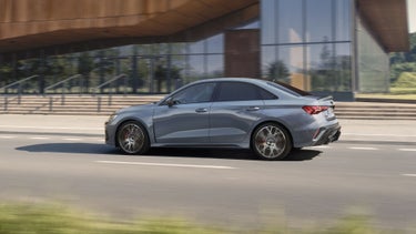 Die RS 3 Limousine von der Seite fährt auf einer urbanen Straße, im Hintergrund ein modernes Bürogebäude.