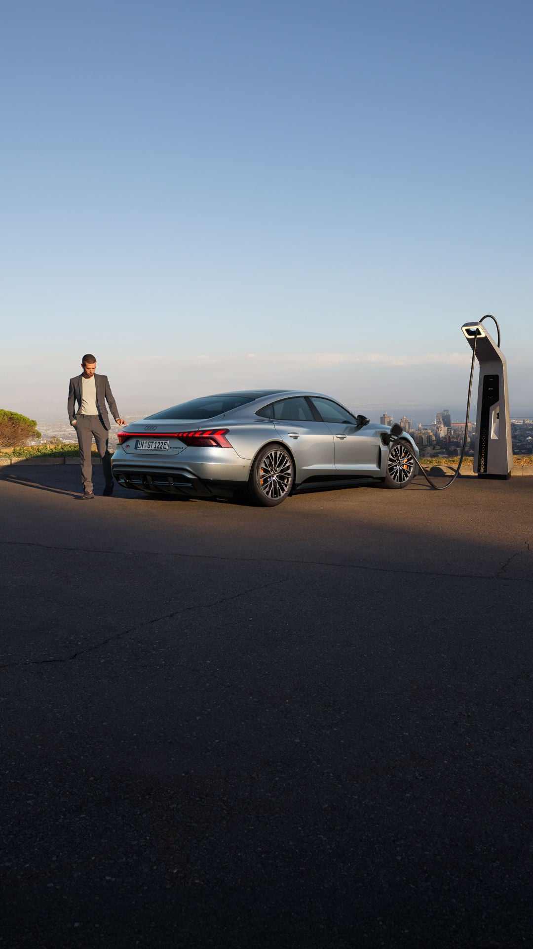 Ein silberner Audi S e-tron GT lädt an einer Ladestation mit Blick auf eine Stadt. Ein Mann im Anzug geht neben dem Auto entlang.