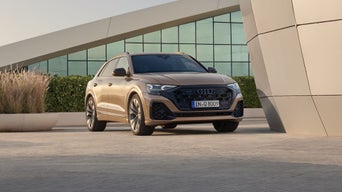 Der Q8 SUV Der Q8 SUV schräg von vorn, neben einem futuristischen Gebäude parkend. Den Parkplatz begrenzt eine grüne Hecke, dahinter die Fensterfront eines weiteren Gebäudes.