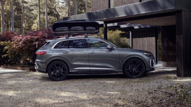 Der Q5 SUV ist mit einer Dachbox ausgestattet und steht seitlich vor einem Haus im Wald.