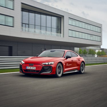 Roter Audi RS e-tron GT performance auf Teststrecke vor modernem Gebäudekomplex in Fahrt.