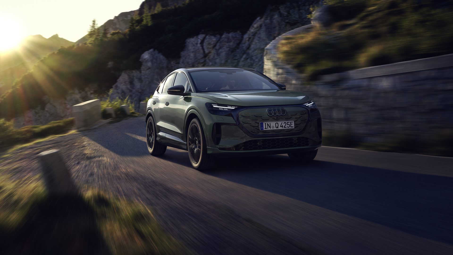 Der Q4 Sportback e-tron bei der Fahrt auf einer Bergstraße. Sonnenstrahlen von links oben beleuchten das Bild, das Fahrzeug wirft Schatten nach vorne.