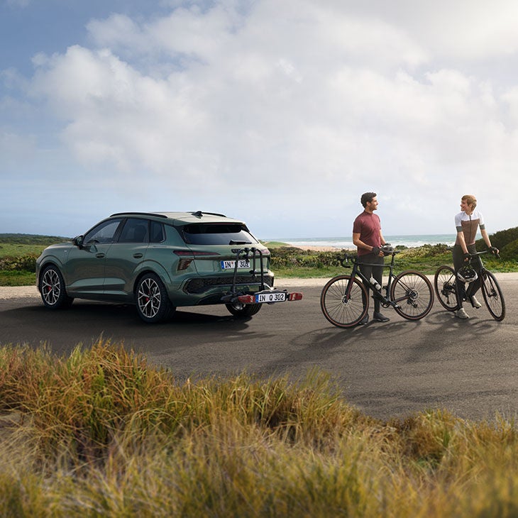 Deux hommes se tiennent à côté d'une Q3 verte équipée d'un porte-vélos fixé sur l'attelage, leurs vélos à la main.