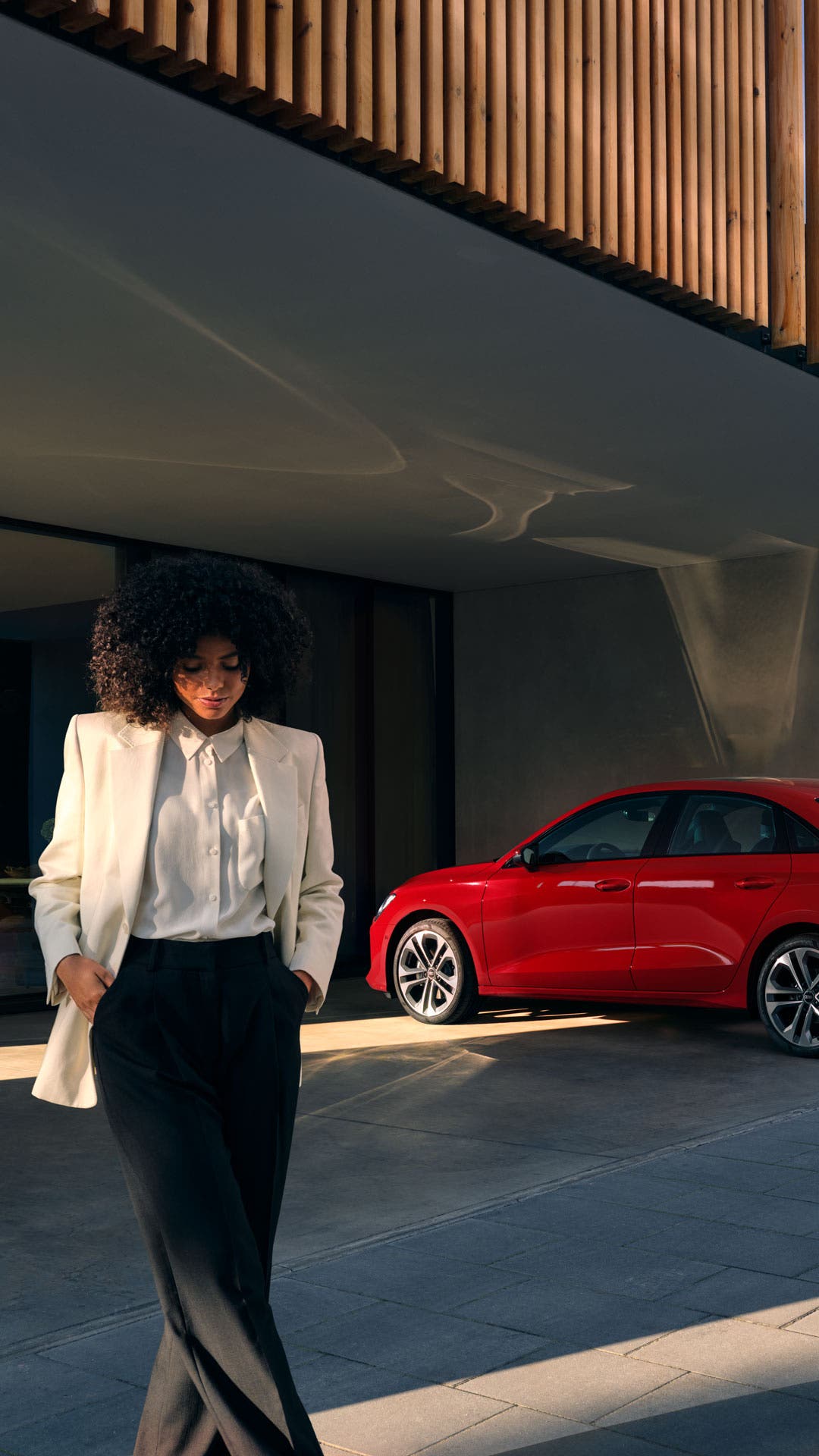 Eine Frau in einem weißen Blazer schaut nach unten, im Hintergrund ein modernes Haus und ein roter Audi.