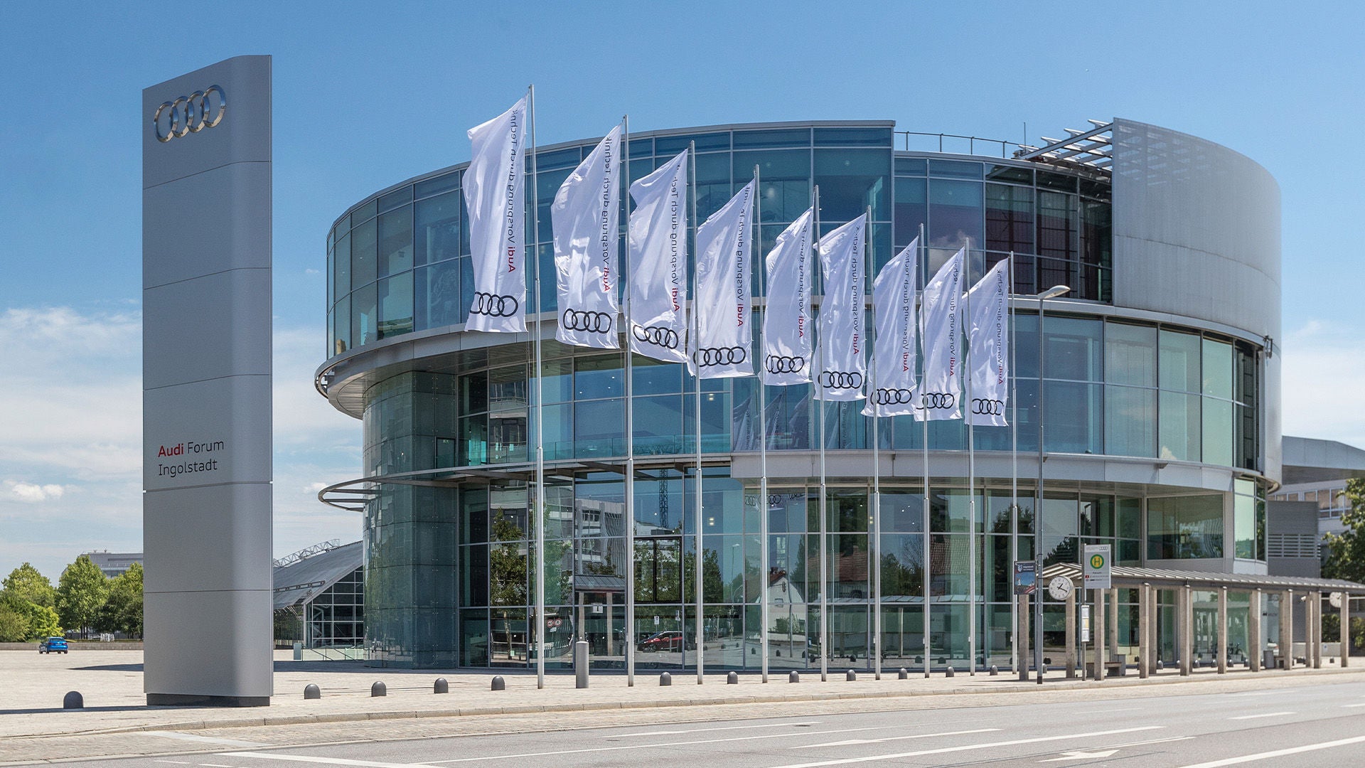 Modernes Glasgebäude des Audi Forum Ingolstadt mit Audi-Flaggen und großem Logoschild vor blauem Himmel.