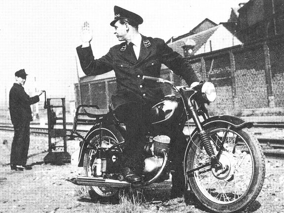 Frontale Schwarz-weiß-Aufnahme von einem Mann in Uniform auf einer DKW RT 125 W, der sich umdreht und einem anderen Mann zuwinkt