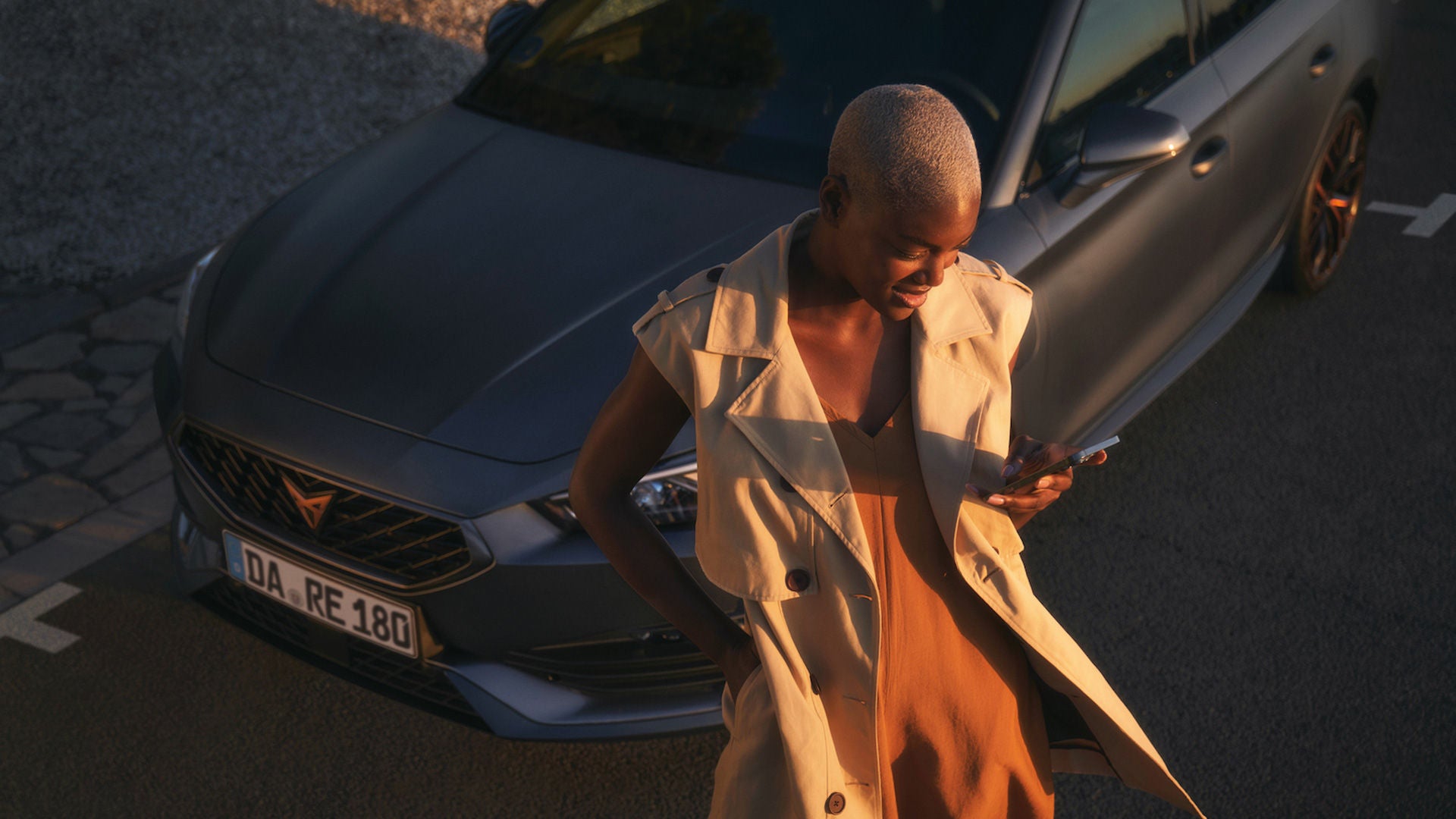 Farbige Frau mit kurzen bonden Haaren steht vor einem grauen CUPRA Modell und schaut auf ihr Smartphone