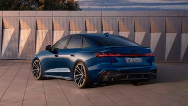 The dark blue S5 Saloon is parked at an angle in front of a building facade and is illuminated by sunlight.
