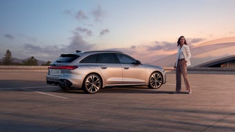 Eine Frau steht neben einem silbernen Audi Modell bei Sonnenuntergang und guckt in die Ferne.