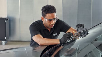 Mechaniker mit Schutzbrille repariert Windschutzscheibe eines Audi mit Spezialwerkzeug in der Werkstatt.