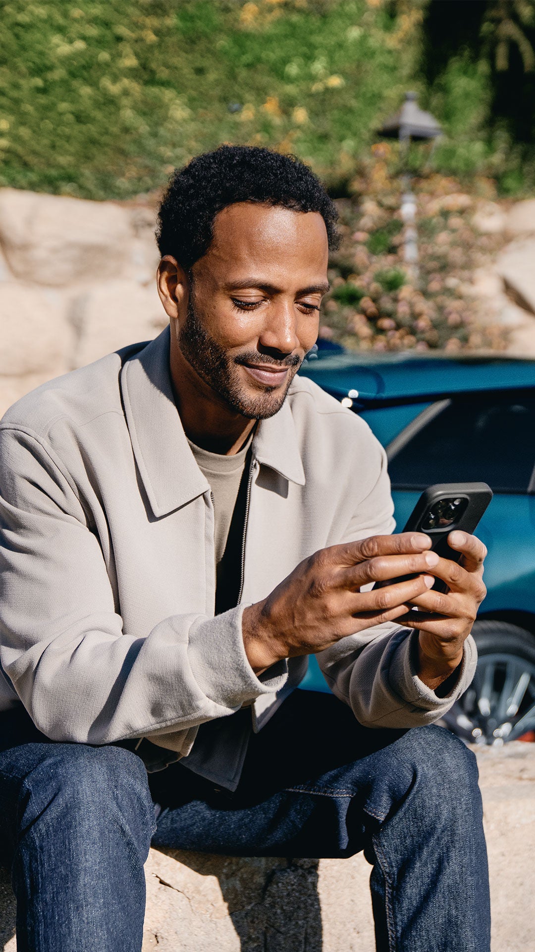 Un homme est assis sur un mur et est occupé par son téléphone portable. Derrière lui se trouve une Audi bleue.
