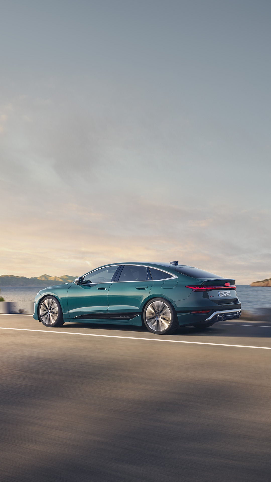 Ein grüner Audi S6 Sportback e-tron fährt auf einer Küstenstraße, Meer und Berge im Hintergrund.