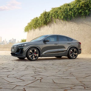 Grey Audi parked on a textured stone surface, with city skyline in the background. Sunlight highlights its modern design. Greenery tops a nearby wall.
