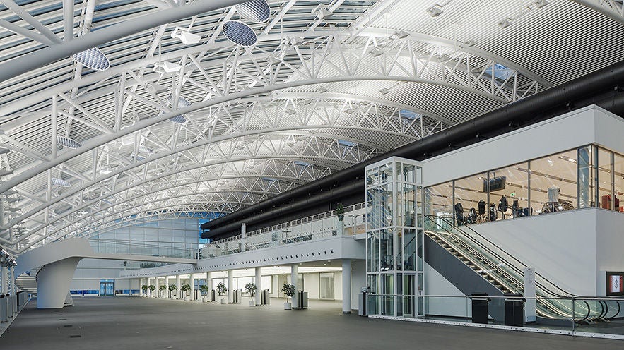 Helle Eingangshalle mit gewölbter Stahlkonstruktion, gläsernen Galerien, Rolltreppe und Aufzügen.