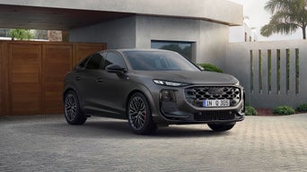 A matte black Audi Q3 parked in front of a modern house with palm trees and wooden garage doors in daylight.