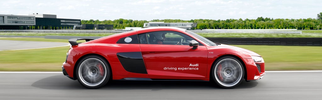 Roter Audi R8 Coupé V10 performance quattro im Seitenprofil auf Rennstrecke, Fahrer mit Helm; weißer Schriftzug „Audi driving experience“ auf der Tür, im Hintergrund ein Audi Gebäude.
