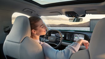 Frau sitzt entspannt mit geschlossenen Augen im Audi SUV Q6 e-tron, mit Blick auf Meer und Sonnenuntergang durch die Frontscheibe.