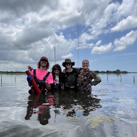 Four people in waders smiling in shallow water under a cloudy sky.