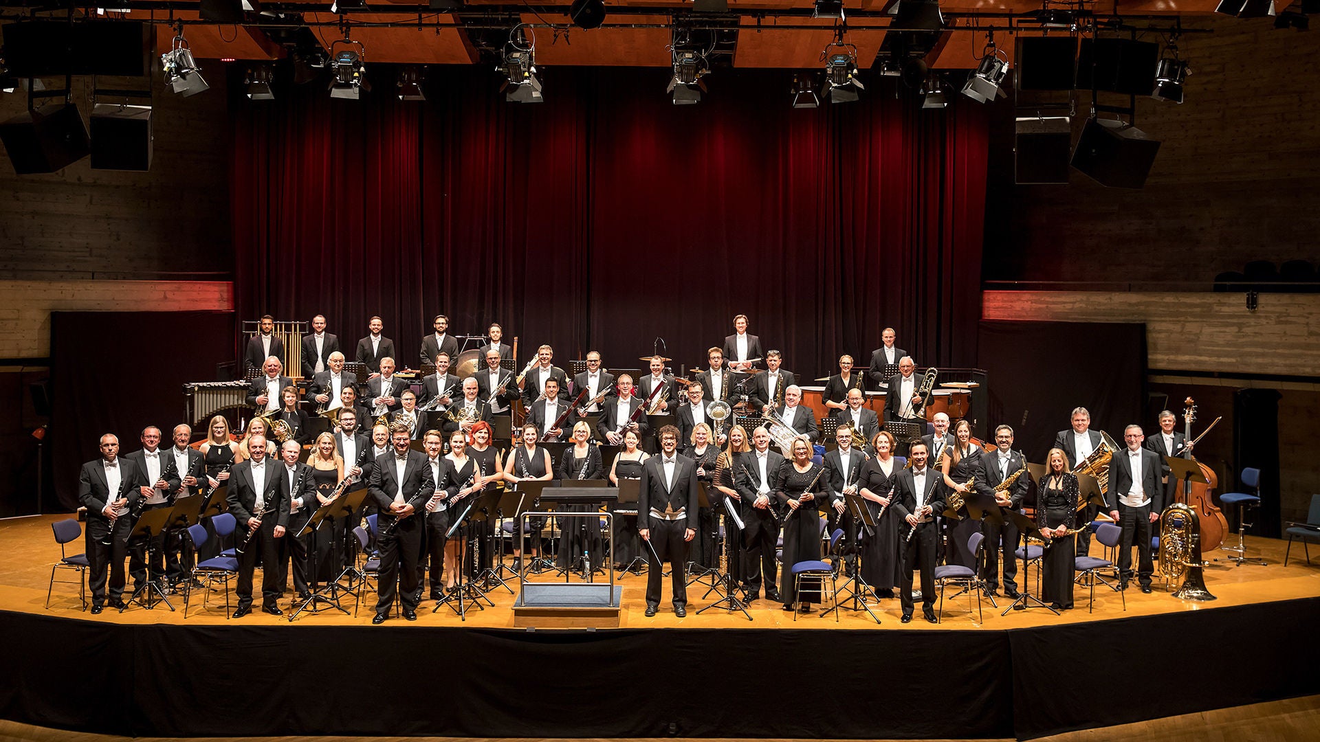 Großes Orchester in Abendgarderobe posiert auf einer Konzertbühne mit Instrumenten vor rotem Vorhang.