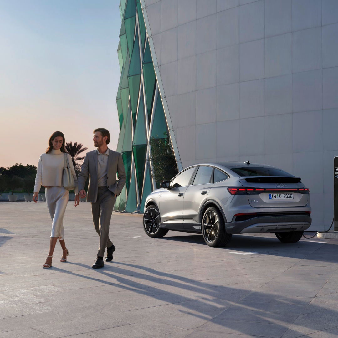 A stylish electric SUV parked next to a modern building, with a couple walking in the foreground against a serene sunset backdrop.