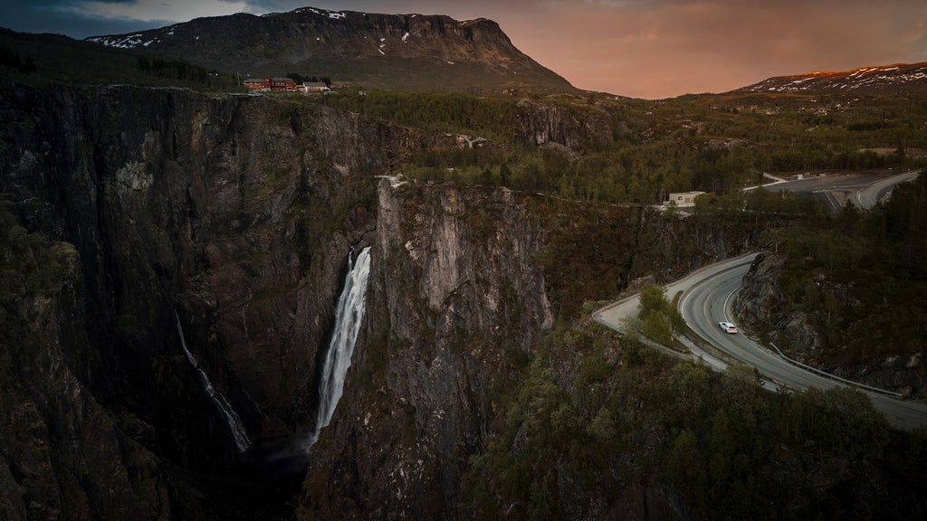 Panoramic view of the mighty mountain ranges in western Norway, looking towards the Vøringsfossen waterfall and winding road, with the Audi A6 Sportback e-tron performance visible in the distance.