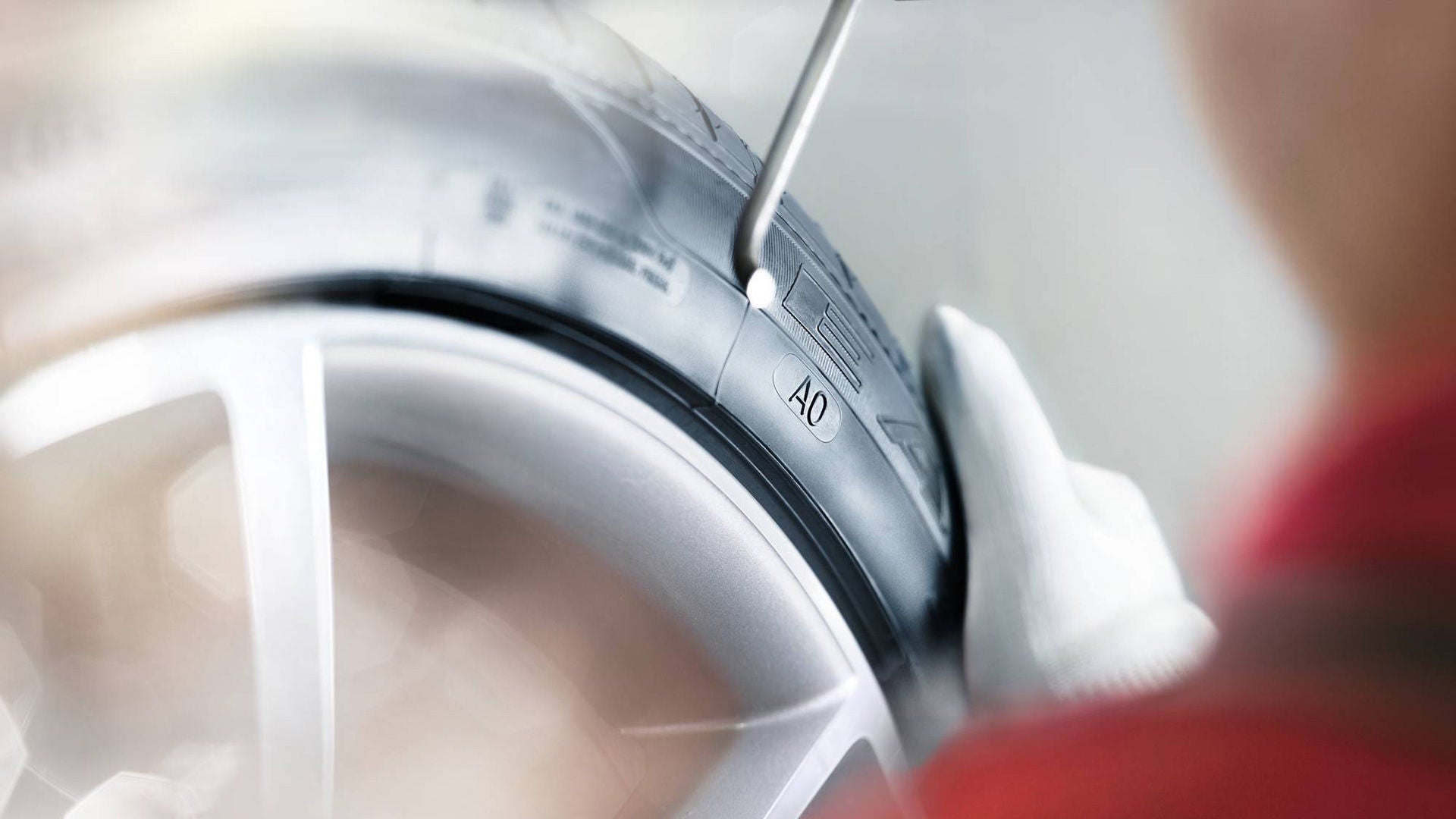 Nahaufnahme eines Autoreifens mit AO-Kennung, eine Hand mit Handschuh prüft das Profil mit einem silbernen Messgerät.