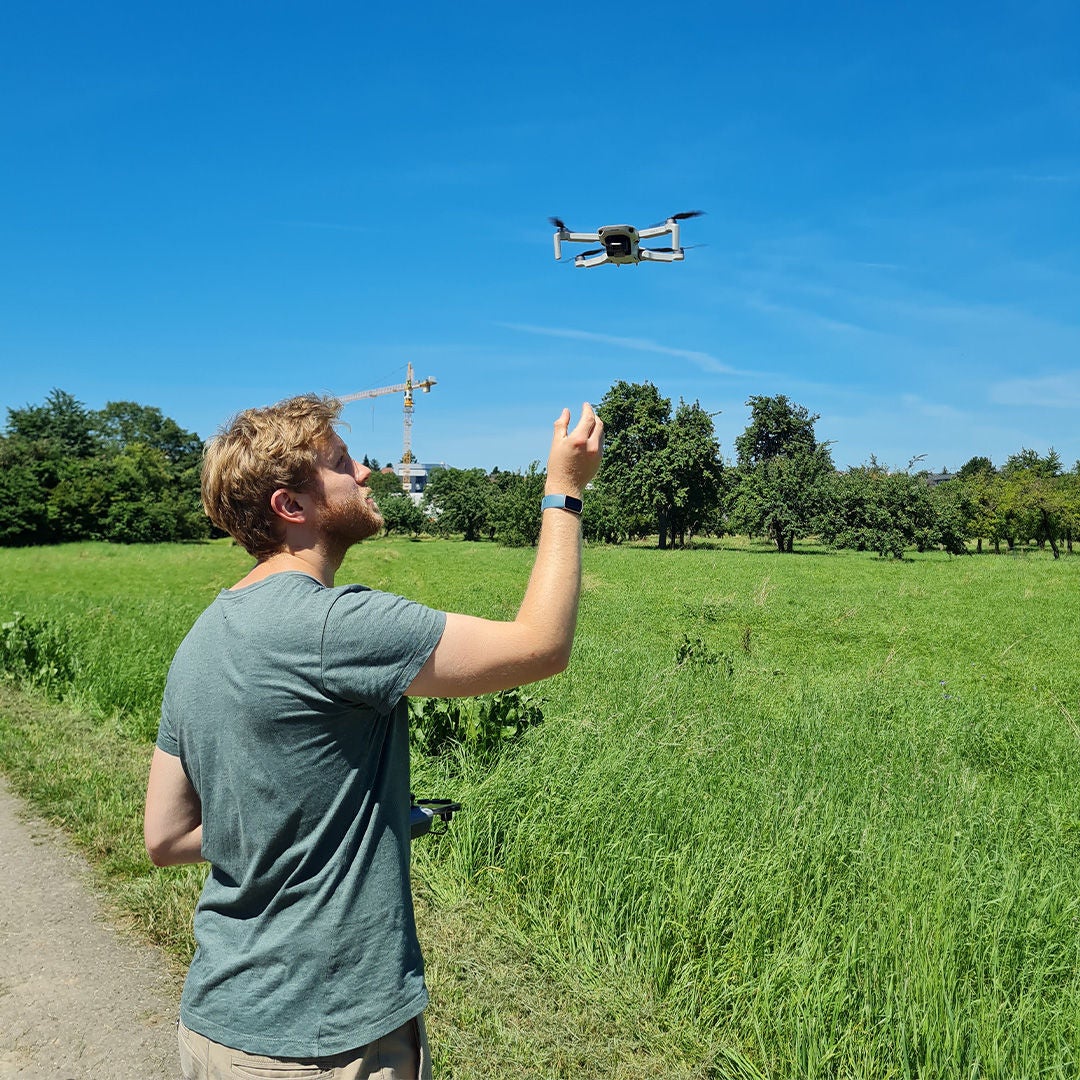 In sunny weather, a man flies a drone over a green meadow with fruit trees and a construction crane in the background.