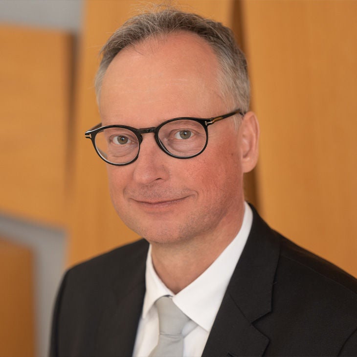 Porträt eines Mannes mit Brille im Anzug vor hellem Hintergrund, er lächelt in die Kamera.