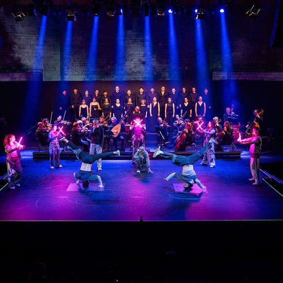 Chor und Orchester begleiten eine Bühnenperformance mit Tänzerinnen und Tänzern in blauem und violettem Licht.