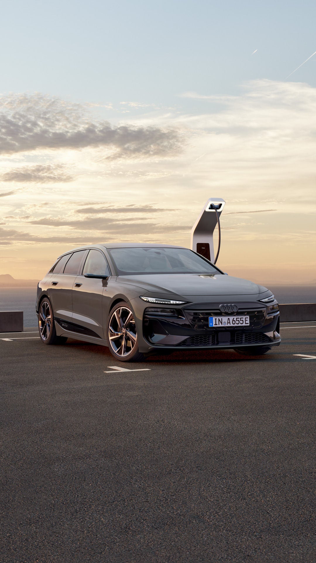Der A6 Avant e-tron steht rückwärts auf einem Parkplatz und ist dabei an eine Ladestation angeschlossen. Im Hintergrund sind das Meer, sowie Berge zu erkennen.&nbsp;